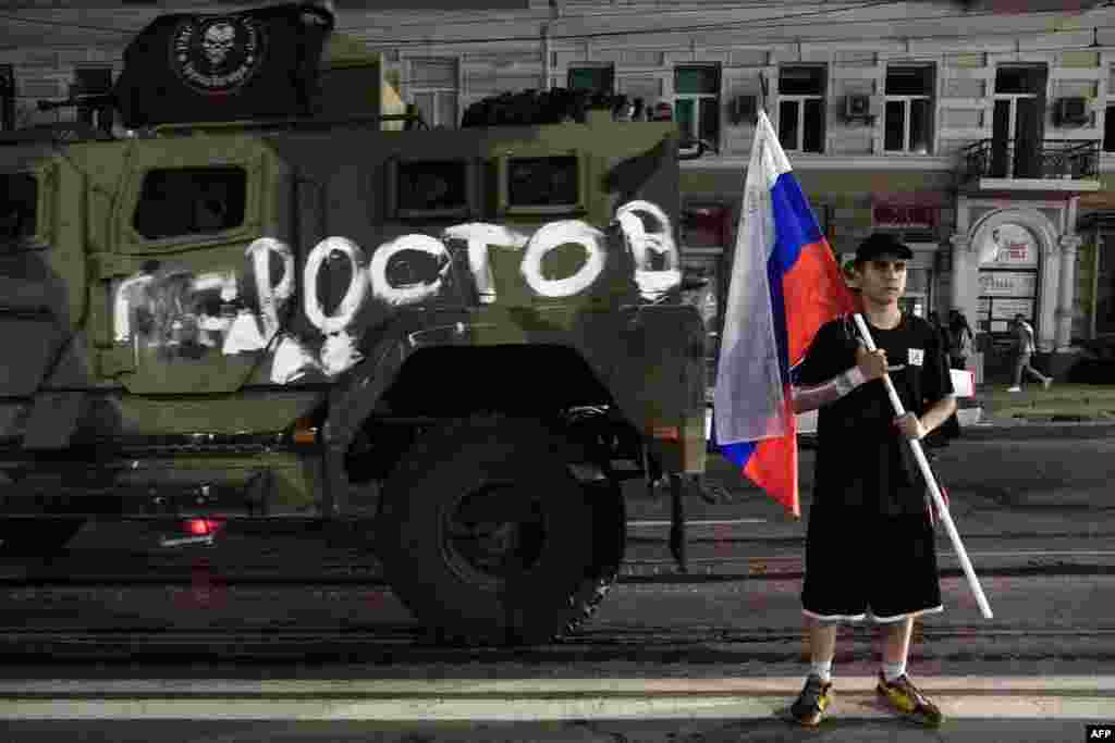 Бүйіріне "Ростов" деп жазылған, "Вагнер" жалдамалы сарбаздар тобының әскери техникасының қасында Ресей мемлекеттік туын ұстап тұрған жас жігіт.